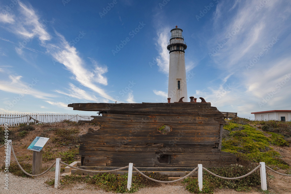 Obraz premium Scenic view of a lighthouse against a blue sky with an old wooden structure nearby. Likely located on the Californian coast, possibly along the 17 mile drive near Pebble Beach.