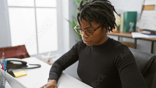 Relaxed yet serious, young black woman with dreadlocks nonchalantly working on laptop in office, giving us a simple, natural look with an earnest expression. photo