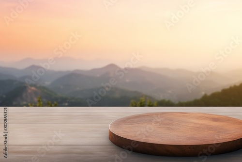 An empty wooden table with a round wooden cutting board on top of it. The table is placed in a beautiful outdoor setting with mountains in the background. The sun is setting and the sky is a warm, ora