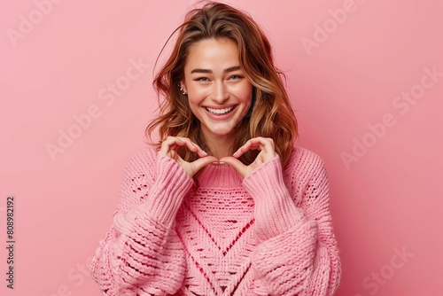 smiling woman making heart shape with hands pink sweater on matching studio background © Lucija