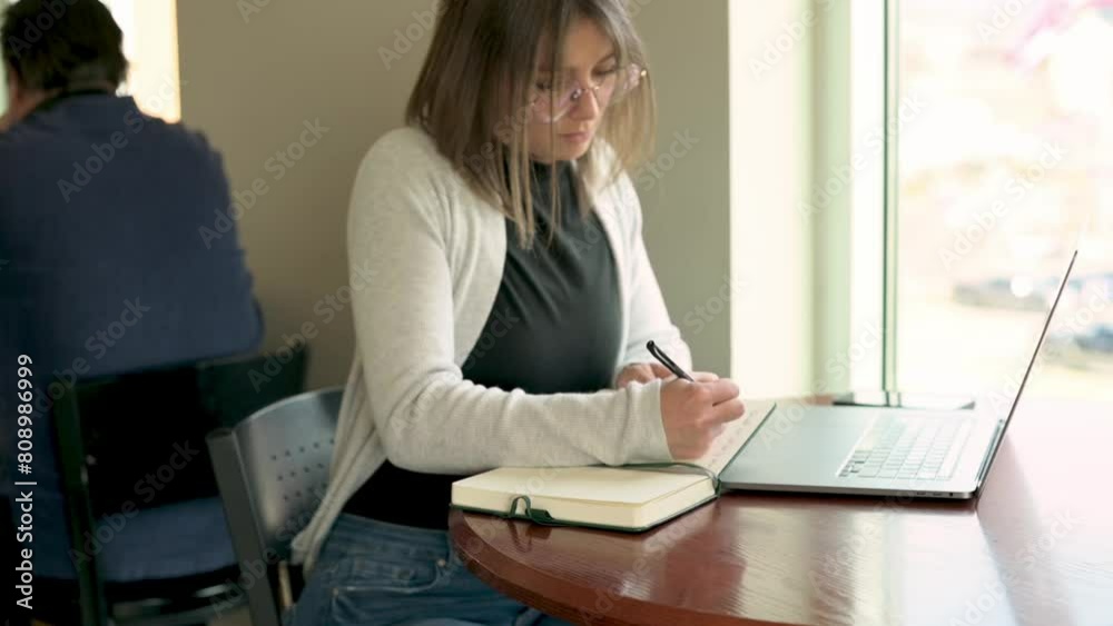 This image portrays a businesswoman deeply engaged in writing notes in her notebook while her laptop stands open beside her, illustrating the integration of analog and digital work methods This image portrays a businesswoman deeply engaged in writing notes in her notebook while her laptop stands open beside her, illustrating the integration of analog and digital work methods