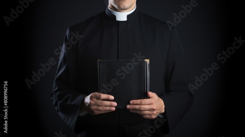 Catholic priest in black robe holds Holy Bible standing in old church closeup. Senior pastor with book prepares for service in wooden building. Man of God prays during ceremony in country church photo
