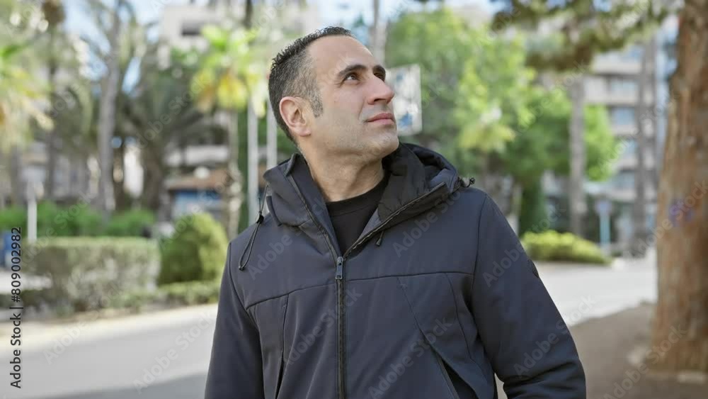 Pensive hispanic man looking upwards in an urban park setting with blurred trees and buildings in the background. Pensive hispanic man looking upwards in an urban park setting with blurred trees and buildings in the background.