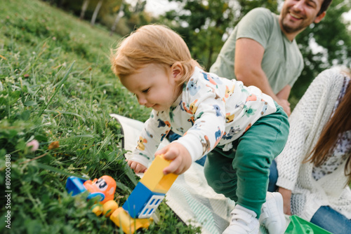 Mother, father, son walking in park. Dad, mom, child 2 year old play constructor game sitting in green grass on blanket sunny summer day. Family with little cute toddler boy playing car toys in garden