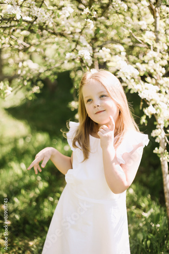 Portrait of little toddler girl in white romantic dress in blossoming garden in spring. Cute beautiful lovely child having fun in a park on a sunny day.