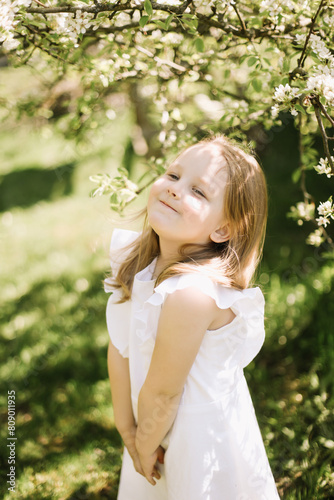 Portrait of little toddler girl in white romantic dress in blossoming garden in spring. Cute beautiful lovely child having fun in a park on a sunny day.