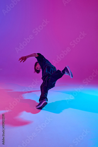 Junior street dancer in dynamic pose dressed in all black in mixed neon light against vibrant gradient background. Concept of sport and hobby, music, fashion and art, movement. Ad