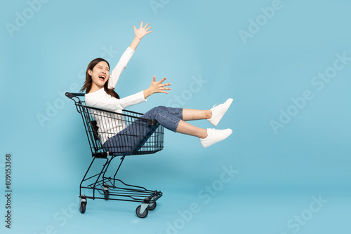 Happy young Asian woman sitting inside of shopping trolley isolated on blue sky background, Wow and surpise concept