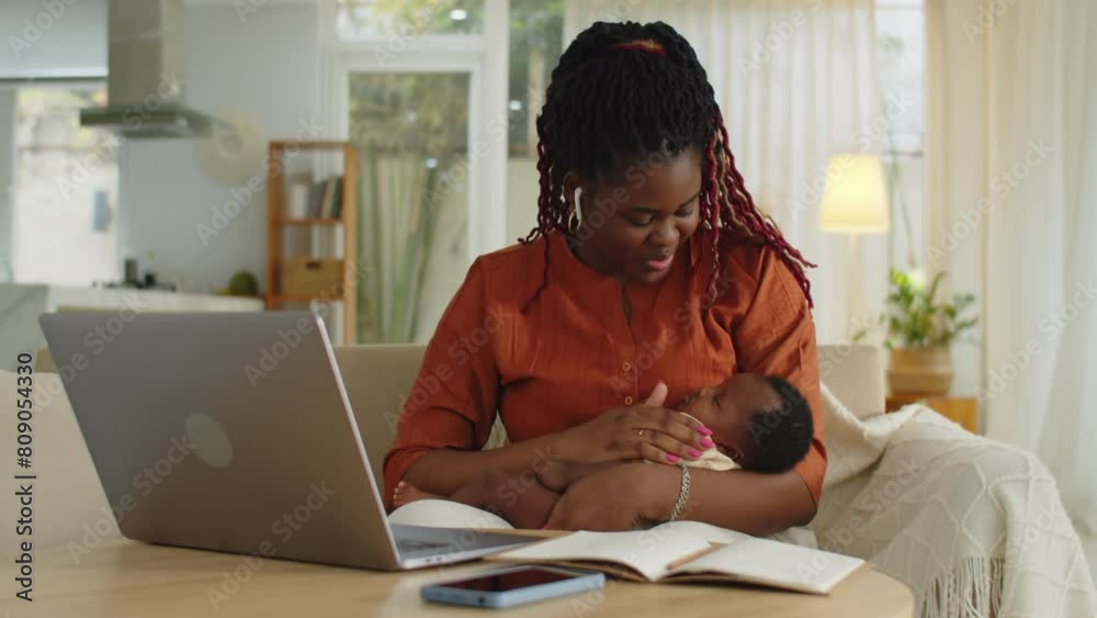 Medium shot of African American businesswoman having online meeting on laptop holding sleeping newborn daughter in hands at home