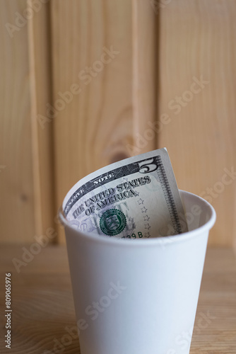 Five dollar bill in a cup on a wooden shelf, payment for coffee, tips in a cafe, donations and contributions, vertical shot photo