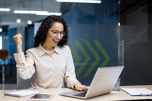 Hispanic business woman celebrating victory success, employee with curly hair inside office reading good news, using laptop at work inside office holding hand up and happy triumph gesture