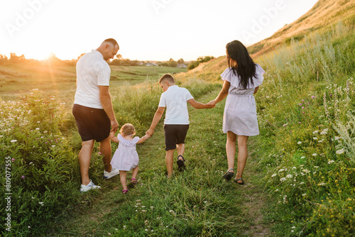 Dad and mom holding hands baby daughter, son walk in mountains at sunset. Family spending time together on a summer day. Children with parents happiness walking in green grass in field. Back view