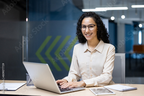 Young beautiful hispanic woman working inside modern office, businesswoman smiling and looking at camera at work using laptop