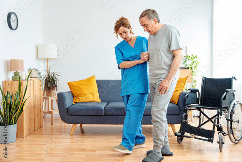 Young caregiver assisting senior man in wheelchair walking indoors - Home health care service and physiotherapy concept photo