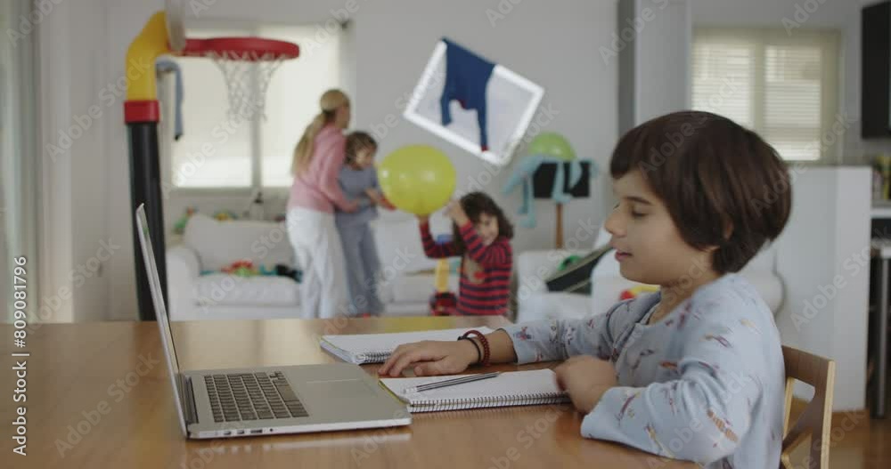 A young boy diligently works on his homework on a laptop at the dining table, as his family members move about in a lively home setting, making noise, playing. A young boy diligently works on his homework on a laptop at the dining table, as his family members move about in a lively home setting, making noise, playing.