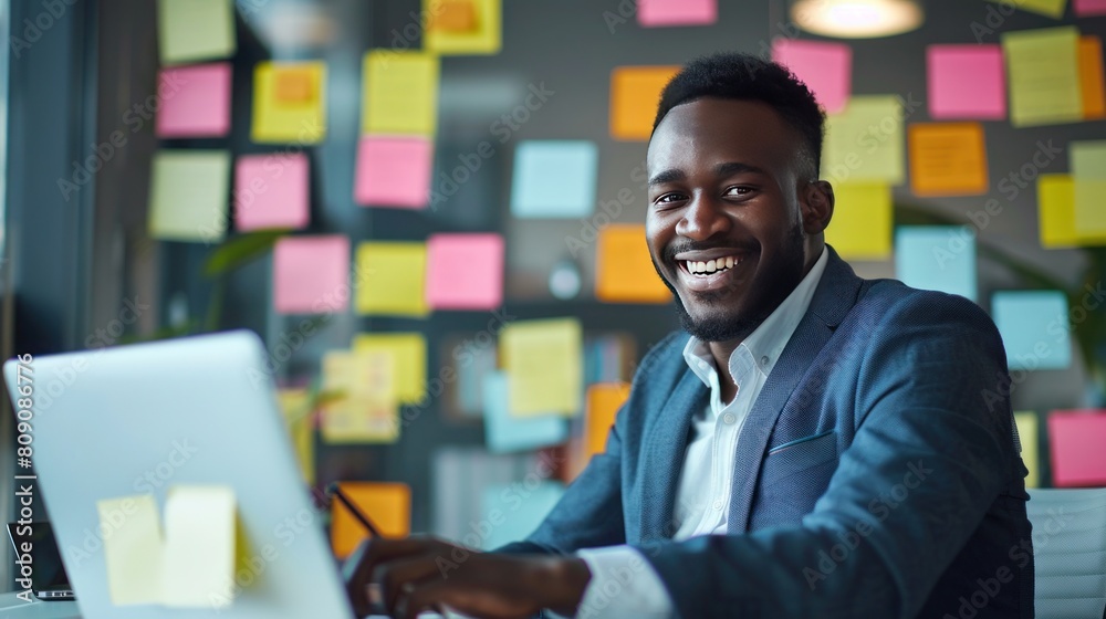 An African businessman is working on a laptop computer with many post-it notes in the background
