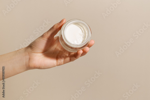 A female hand holds a glass jar with white face cream on a beige background. The concept of beauty, skin care, cosmetology.
