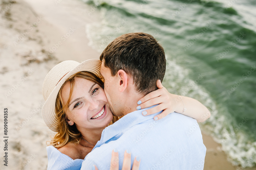 Fototapeta premium Couple in love hugging and kissing on seashore. Male kisses female on cheek on beach ocean and enjoys sunny summer day. Man embraces woman on sand sea. Spending time together. Closeup face. Top view