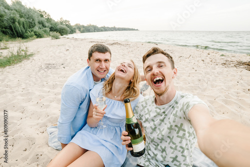 Selfie photo. Friends with champagne relax on picnic in nature spending time together. Men and woman sitting on blanket on the sand sea. Female and males on beach ocean and enjoying sunny summer day. © Serhii