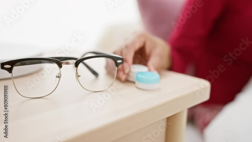 In a bright bedroom, a woman takes lens out of a case on a nightstand with eyeglasses in the foreground. photo