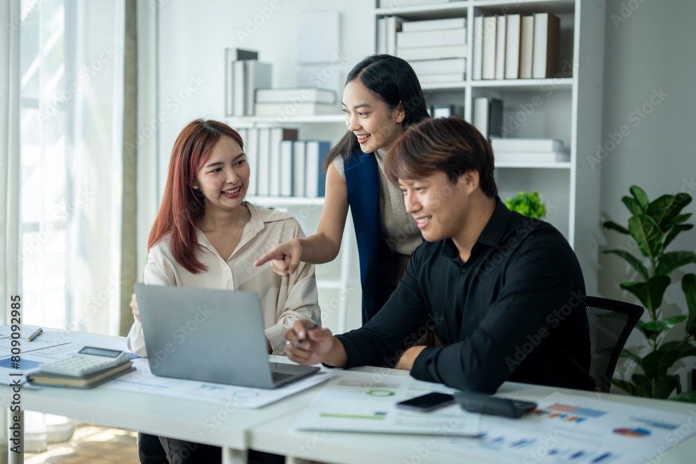 Three people are sitting at a desk with a laptop open. One of them is pointing at the screen. Scene is collaborative and focused Three people are sitting at a desk with a laptop open. One of them is pointing at the screen. Scene is collaborative and focused