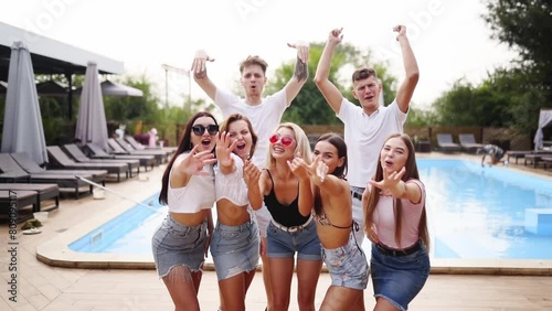 Company of friends posing for common photo at swimming pool with sun loungers before party in sunny summer day. Model men and women waving, shouting, smiling at camera and call to join. photo