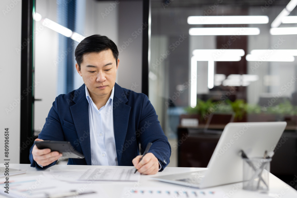 Focused Asian businessman reviewing financial reports while working in a modern office space. Concept of professionalism and diligence. Focused Asian businessman reviewing financial reports while working in a modern office space. Concept of professionalism and diligence.