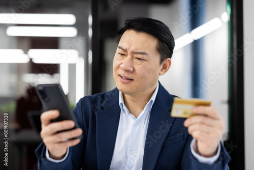 Slika na platnu A middle-aged Asian businessman looks confused and concerned while comparing his credit card to his smartphone in an office setting