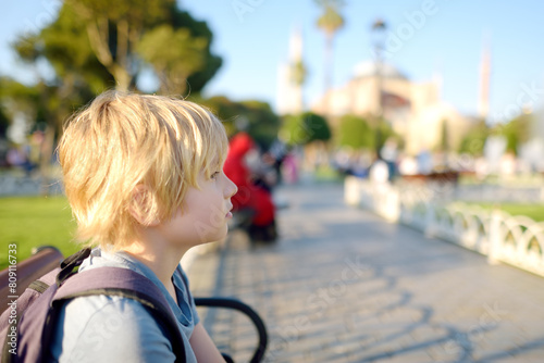 Cute blonde preteen tourist boy sitting on a bench near Sultan Ahmed Mosque district on background of Hagia Sophia Mosque on sunny summer day. Travel, tourism, sightseeing in Istanbul, Turkey. photo