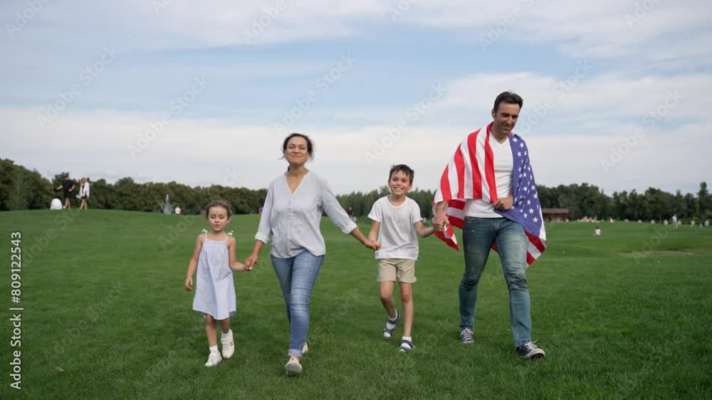 A happy family enjoys leisure time walking in a field with an American flag, surrounded by trees, grass, and a beautiful sky with clouds A happy family enjoys leisure time walking in a field with an American flag, surrounded by trees, grass, and a beautiful sky with clouds