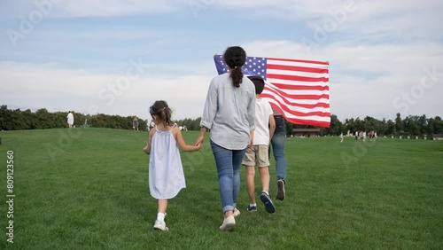 A woman is walking with two children in a grassy meadow, holding an American flag under a cloudy sky. They gesture happily, enjoying leisure time outdoors