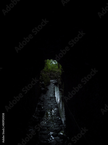 View through exit of tunnel along levada in dense tropical laurel forest at Levada Caldeirao Verde and Caldeirao do Inferno hiking trail, Madeira island, Portugal. Dark black background, copy space photo