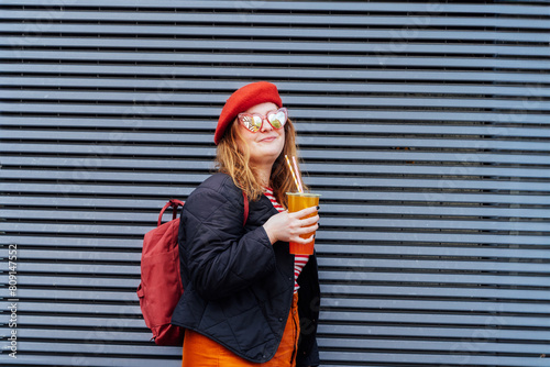 Smiling plus size woman in heart shaped sunglasses and bright clothes drinking sugar flavored tapioca bubble tea near striped urban wall. Happy fashionable hipster overweight girl. Street fashion.