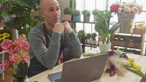 A contemplative bald man with a beard sits at a wooden table in a flower shop, surrounded by arrangements, using a laptop. photo