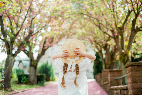Happy pretty girl 6 years old in white dress in straw hat near blooming pink sakura. The child is walking in the park. Spring.