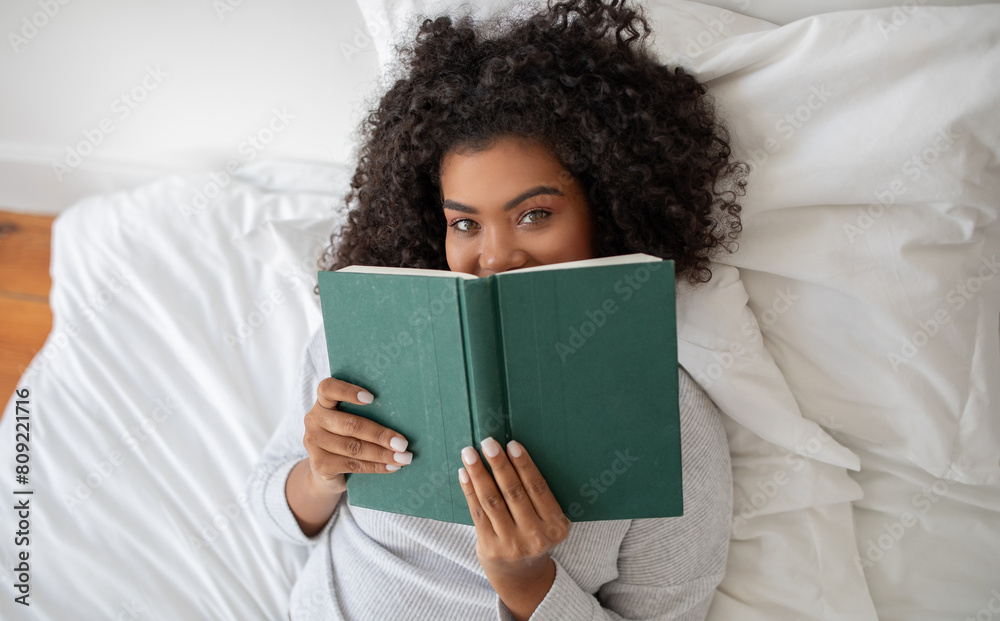 Hispanic woman is laying comfortably in bed, engrossed in a book she is reading. The room is softly lit, top view Hispanic woman is laying comfortably in bed, engrossed in a book she is reading. The room is softly lit, top view