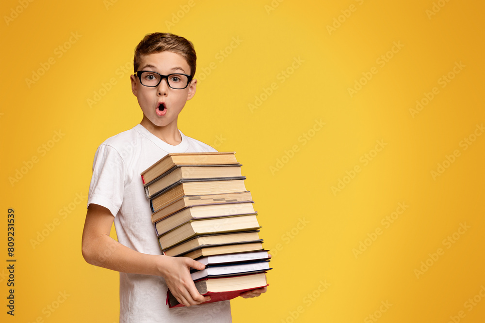 Remote education concept. Shocked schoolboy in glasses holding stack of books, orange panorama background Remote education concept. Shocked schoolboy in glasses holding stack of books, orange panorama background