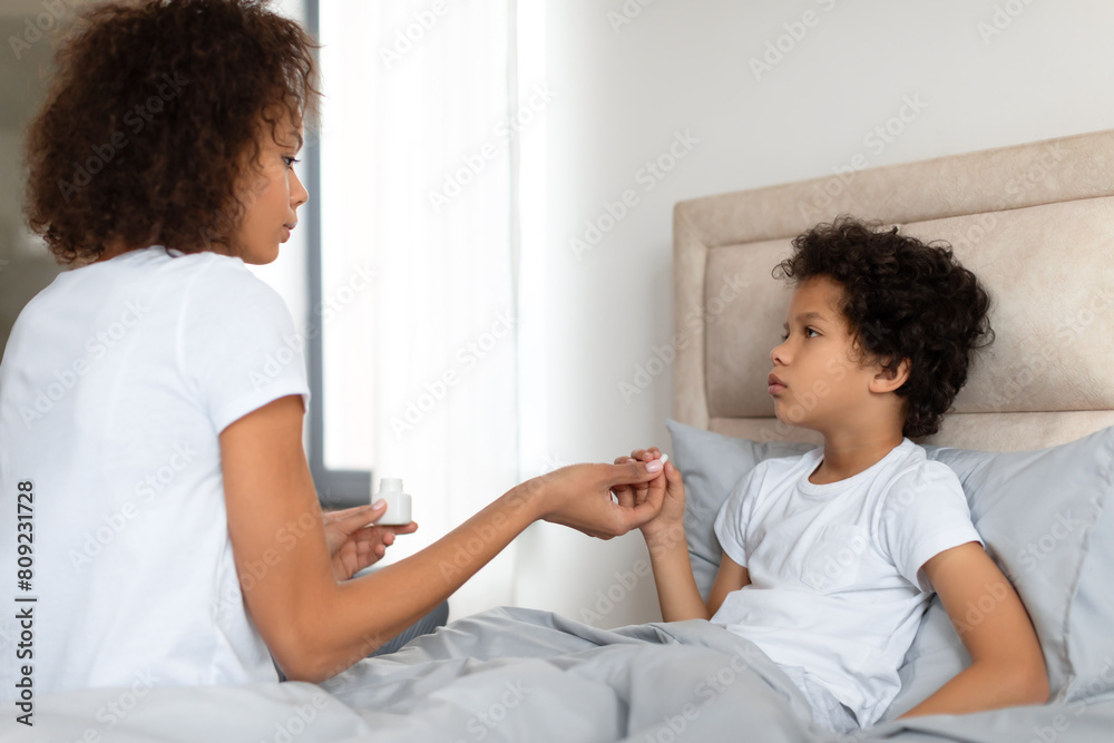Fototapeta premium A concerned African American mother sits on the edge of a bed, gently holding her young child hand as she checks for a fever. The child, looking unwell
