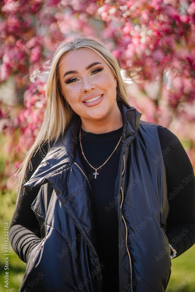a smiling young woman with blonde hair, standing outdoors with a background of blooming pink flowers. a smiling young woman with blonde hair, standing outdoors with a background of blooming pink flowers.