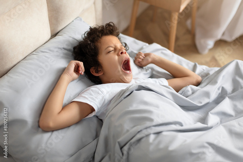 African American young boy is laying in bed, his mouth wide open in a yawn. He appears tired as he stretches his arms out. The background shows a cozy bedroom setting. photo