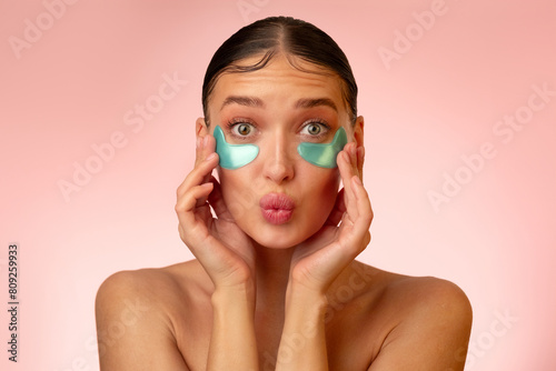 Pretty playful lady wearing eye patches for fresher look, looking at camera and pouting lips over light pink background. Eye skin treatment photo