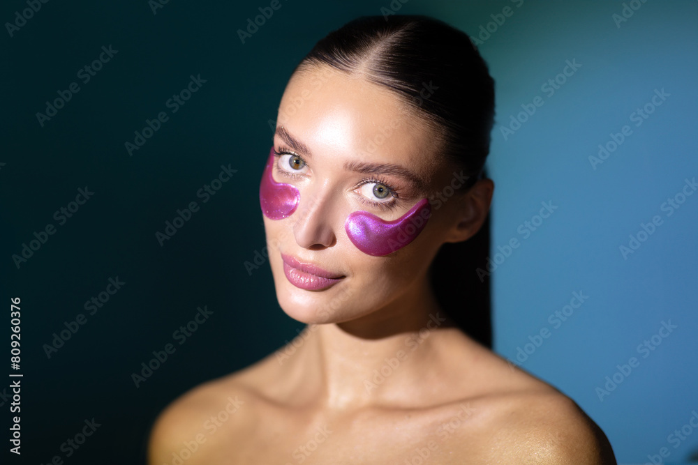 Pretty young woman with under eye patches, enjoy procedures and looking at camera, posing isolated on dark blue background with back light. Skin care, natural beauty, spa treatment