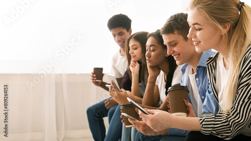 A group of multiethnic teenagers is seated close to one another, each engrossed in their own smartphones. They appear to be in a relaxed social setting photo
