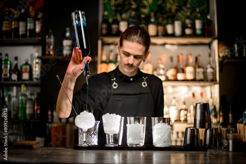 Competent bartender pours a blue drink from a bottle into prepared glasses standing on the bar counter
