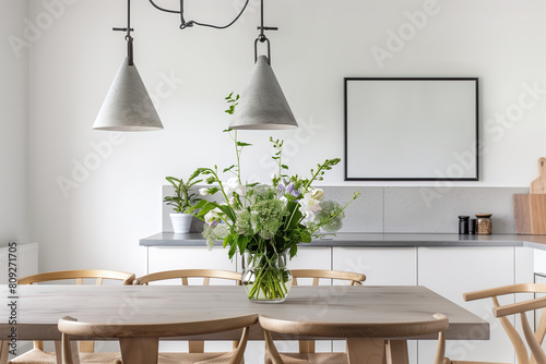 Modern dining room with wooden table, chairs, and concrete pendant lamps over the table decorated by fresh flowers in a vase on grey countertop against white wall with blank frame picture above it.