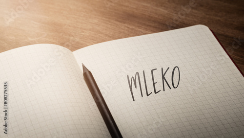  A handwritten inscription "Mleko" on a grille of an open notebook on a wooden countertop, next to a black pencil, lighting of light. (selective focus), translation: milk