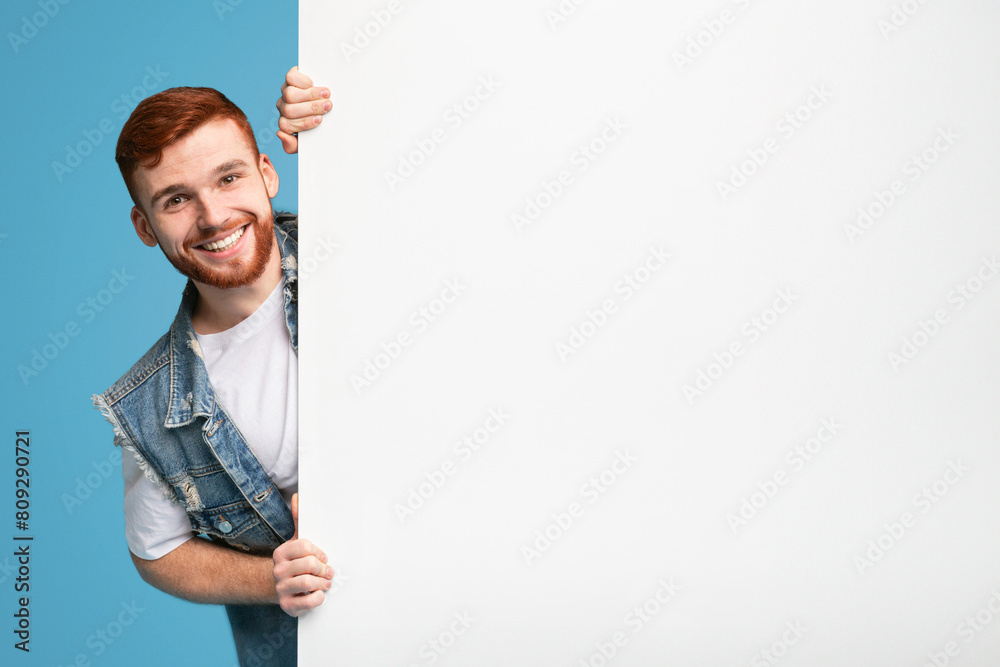 Happy smiling guy looking out of white blank placard ready for advertisement, blue background Happy smiling guy looking out of white blank placard ready for advertisement, blue background