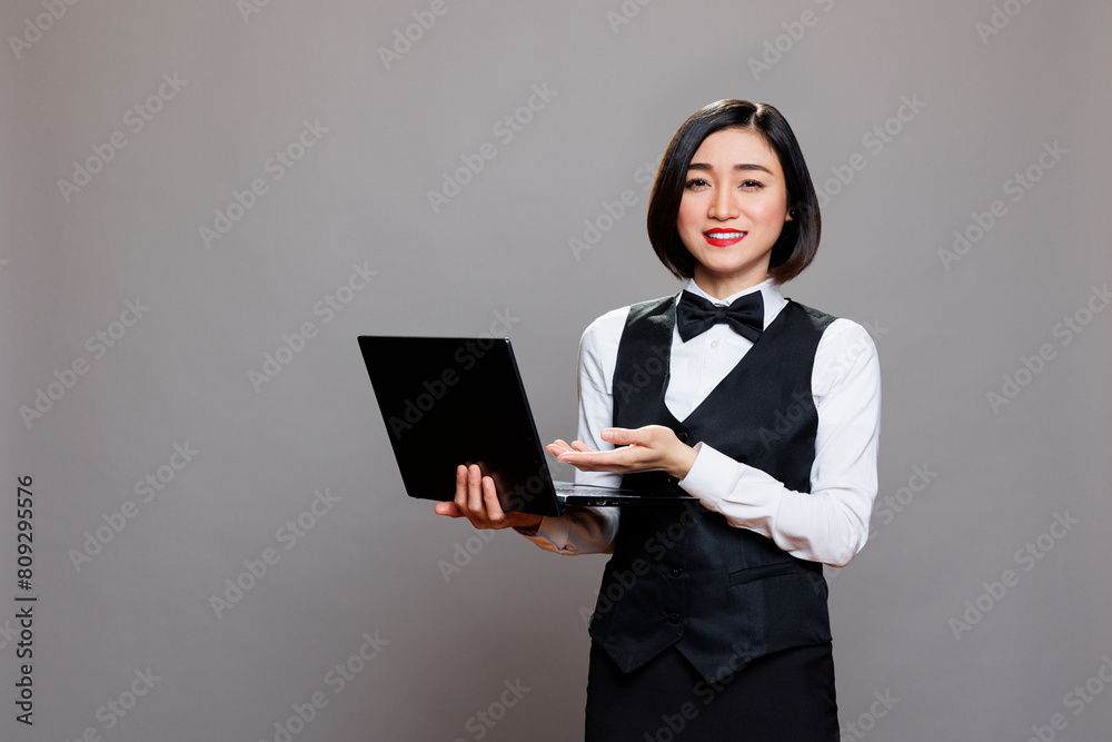 Smiling asian waitress in professional uniform showing laptop and looking at camera. Young attractive receptionist pointing with hand while holding portable computer portrait Smiling asian waitress in professional uniform showing laptop and looking at camera. Young attractive receptionist pointing with hand while holding portable computer portrait