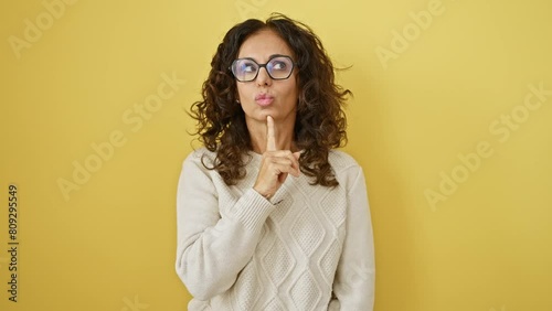 Confident middle-aged hispanic woman in glasses, wonderingly lost in deep thoughts, finger on chin, gazing upwards onto a yellow isolated background