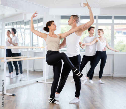 Confident concentrated man, ballet dancer, providing support of graceful female partner in challenging movements, during group rehearsal in vibrant choreography studio